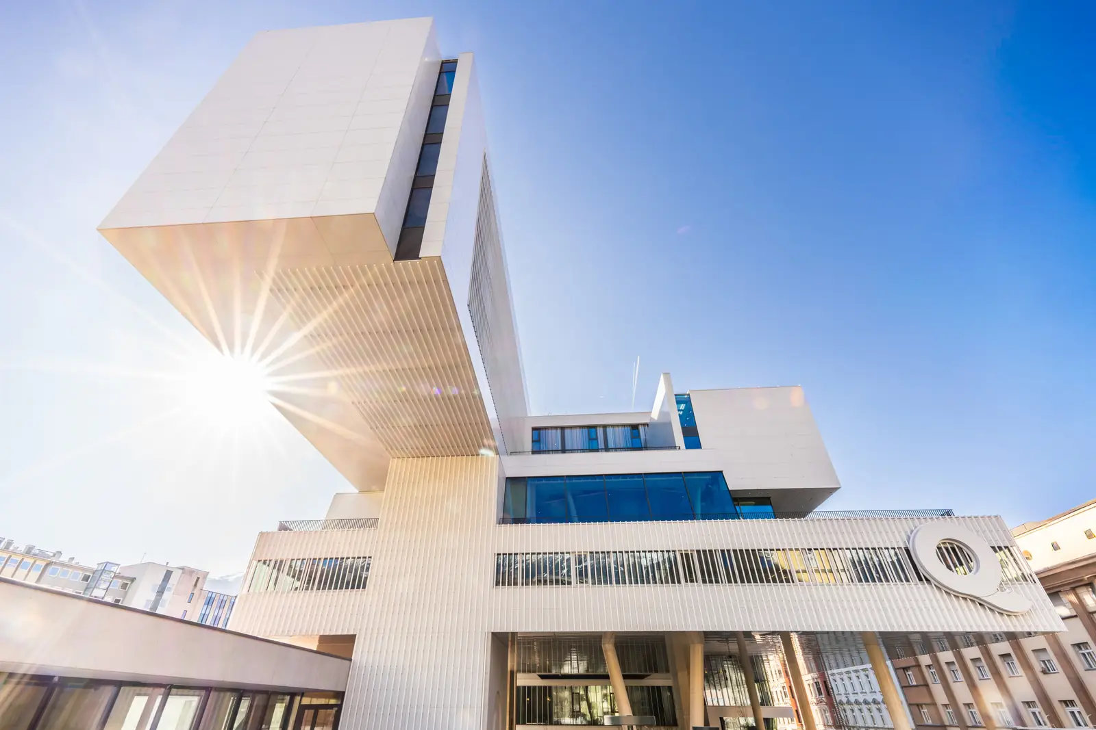 DAS RAIQA in Innsbruck seen from below against the light: the cantilevered ‘hammerhead’ against a blue sky, the sun beside it – venue of the Future Leadership Day Innsbruck