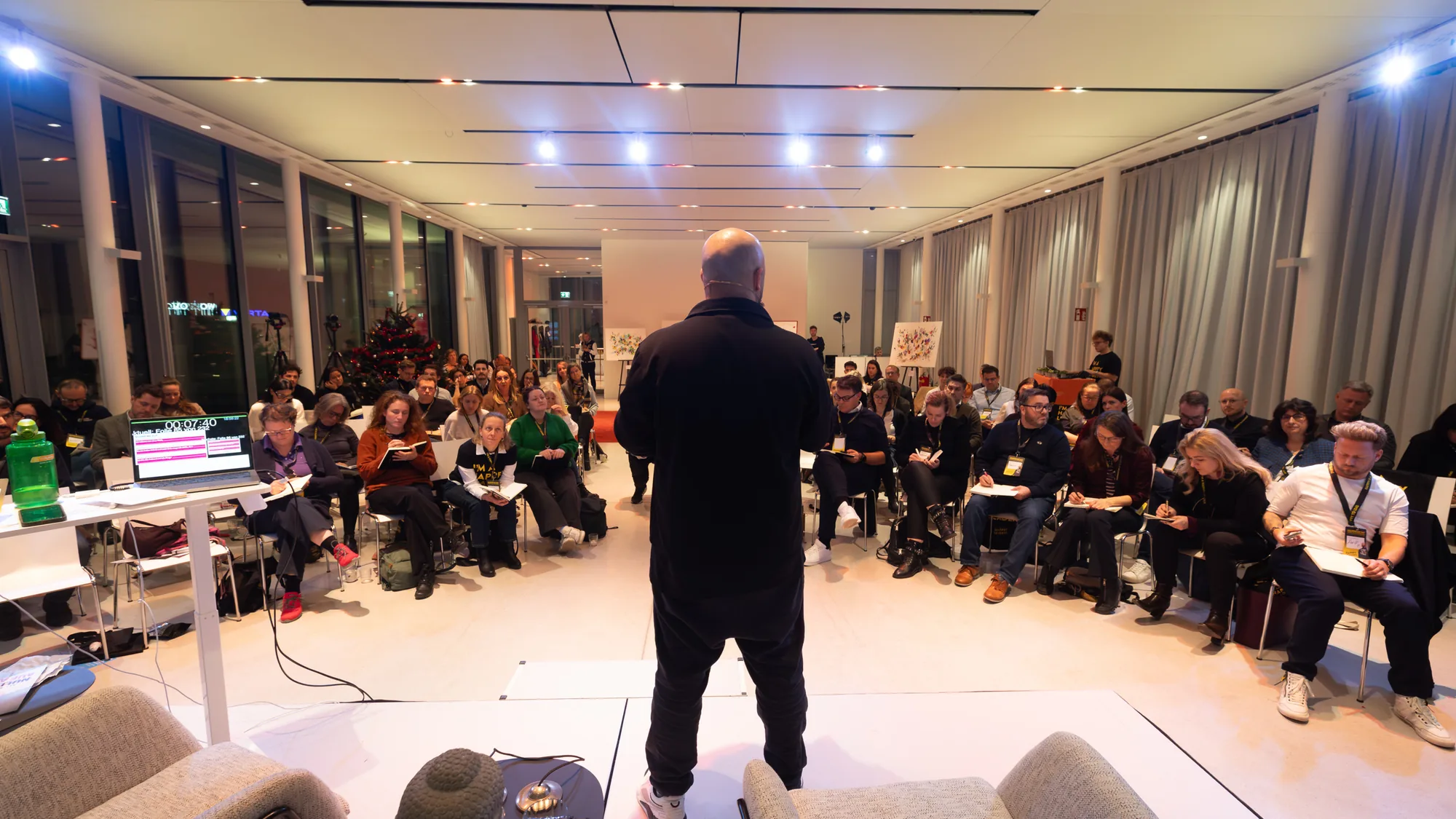 Ali Mahlodji seen from behind in the middle of a large circle of chairs, the participants listening to him