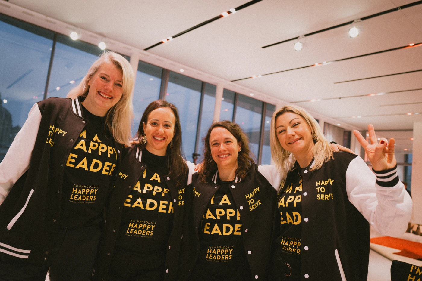 Four women from the Happpy Leaders crew beaming arm in arm into the camera – in “I’m a Happpy Leader” T-shirts and “Happpy to serve” college jackets