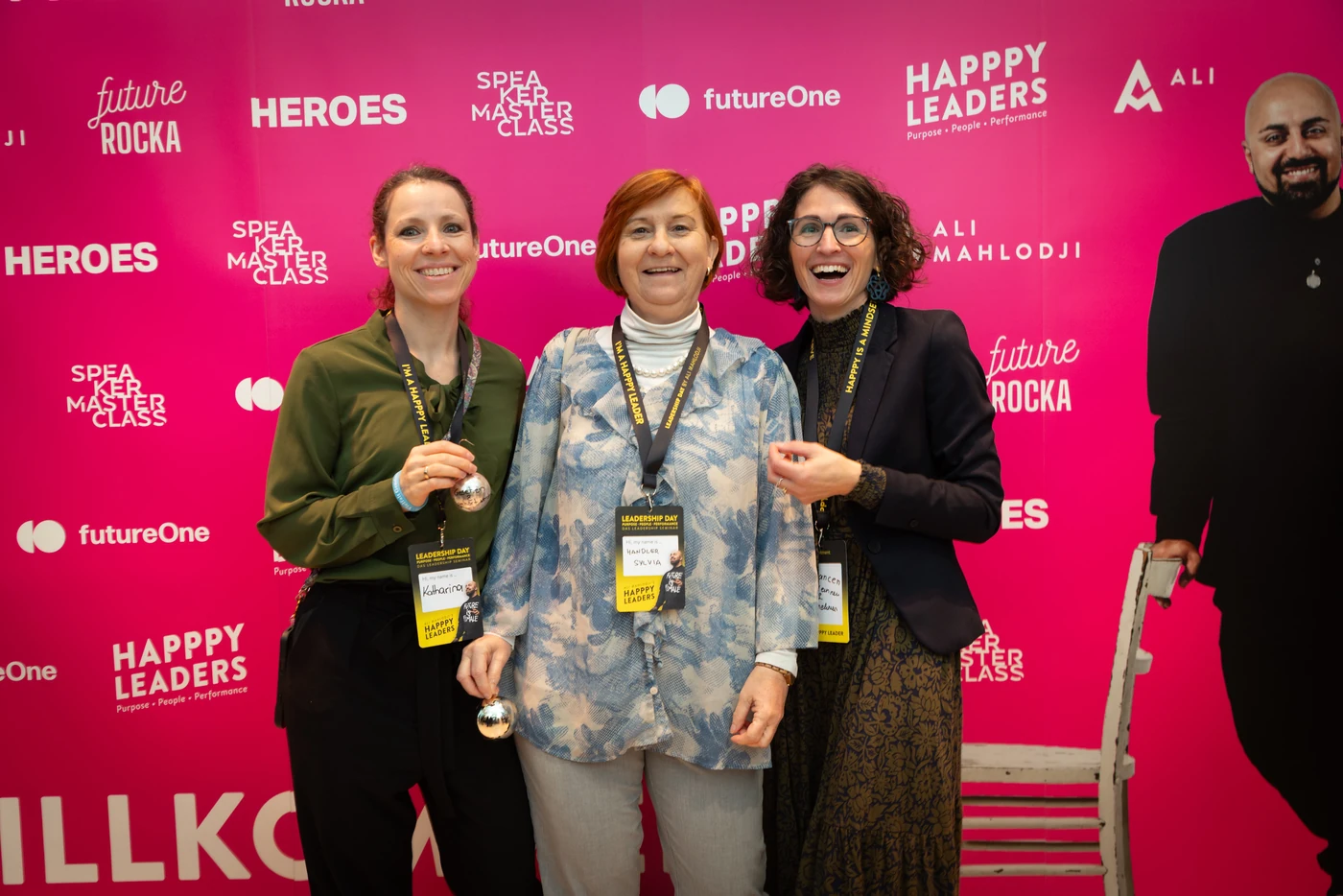 Three participants laughing in front of the pink photo wall with the logos of Happpy Leaders, futureOne and Heroes, two of them holding Christmas baubles, a life-size cut-out of Ali Mahlodji on the right