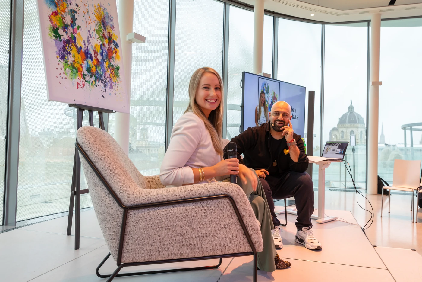 Special guest Lisa Farkas laughing into the camera on stage next to Ali Mahlodji, the dome of a Vienna museum in daylight behind the glass façade