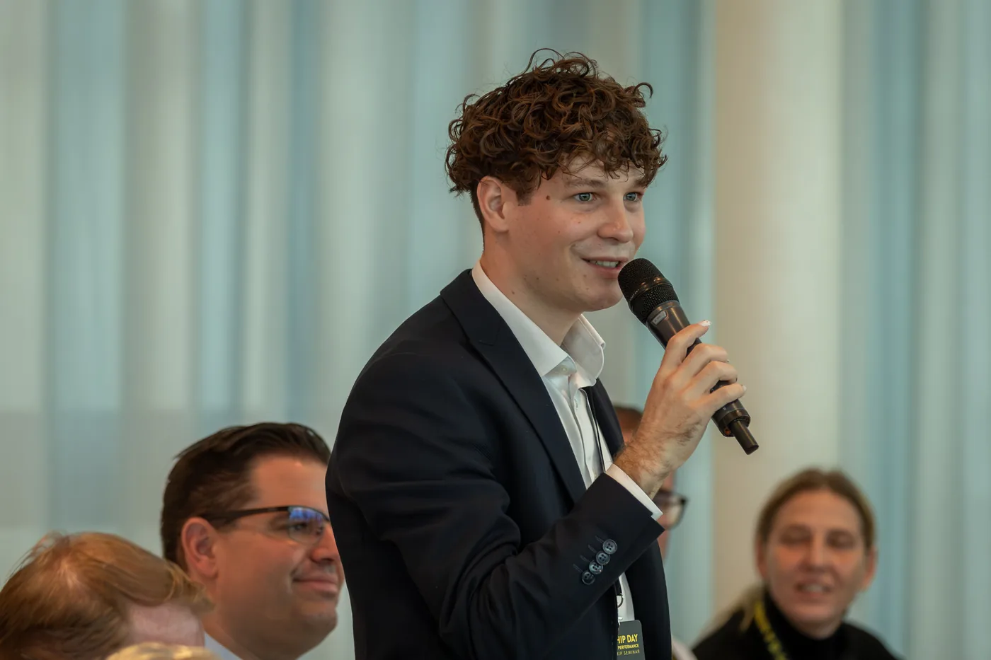 A participant speaking into a microphone to the group, smiling listeners next to him