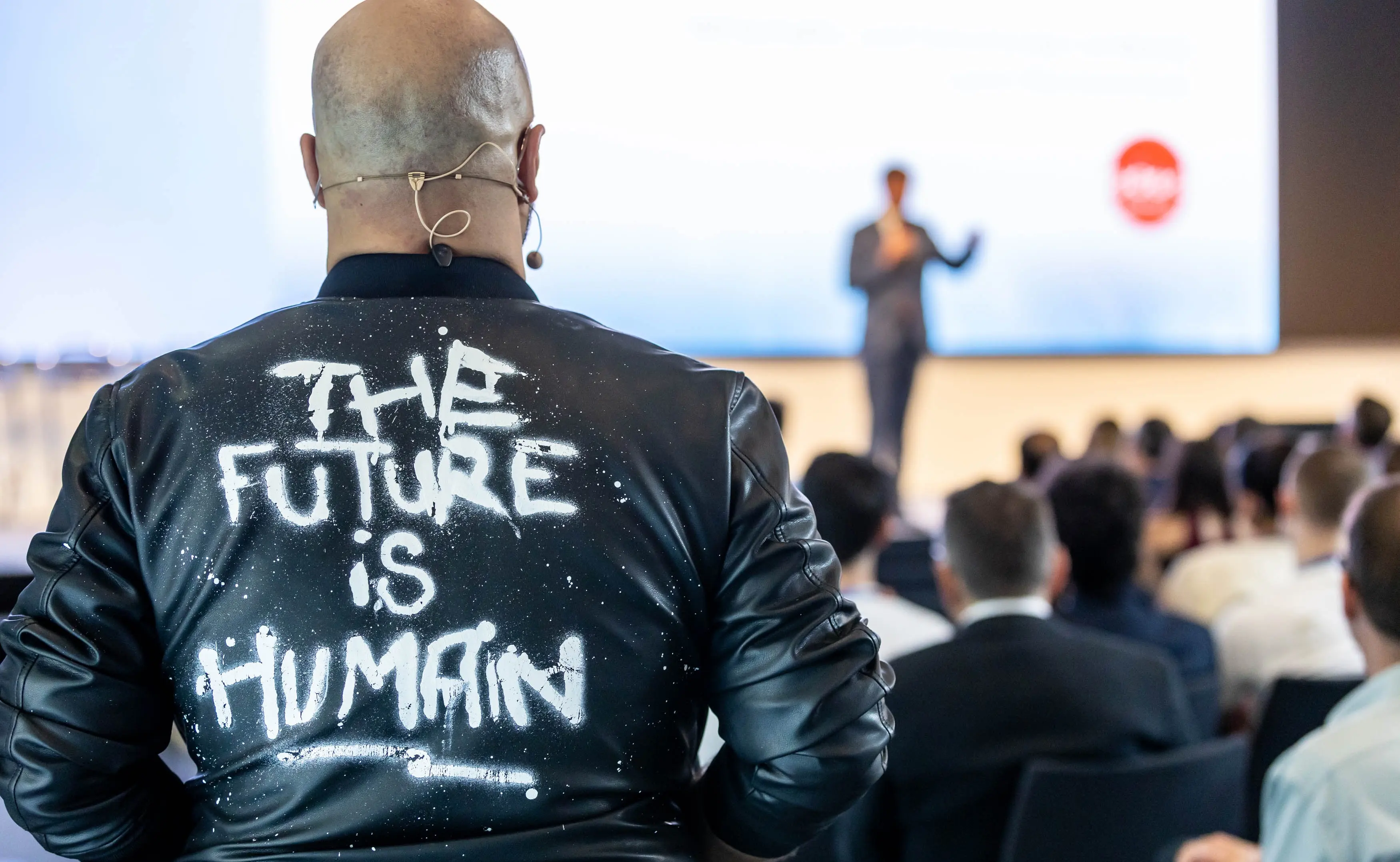 Ali Mahlodji von hinten auf der Bühne, auf der Jacke steht „The future is human“, vor ihm das Publikum