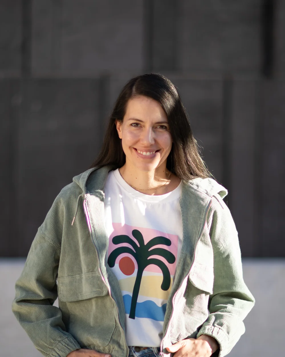 Anna Mahlodji, portrait, smiling in front of a dark stone wall, wearing a palm-tree shirt and a green jacket