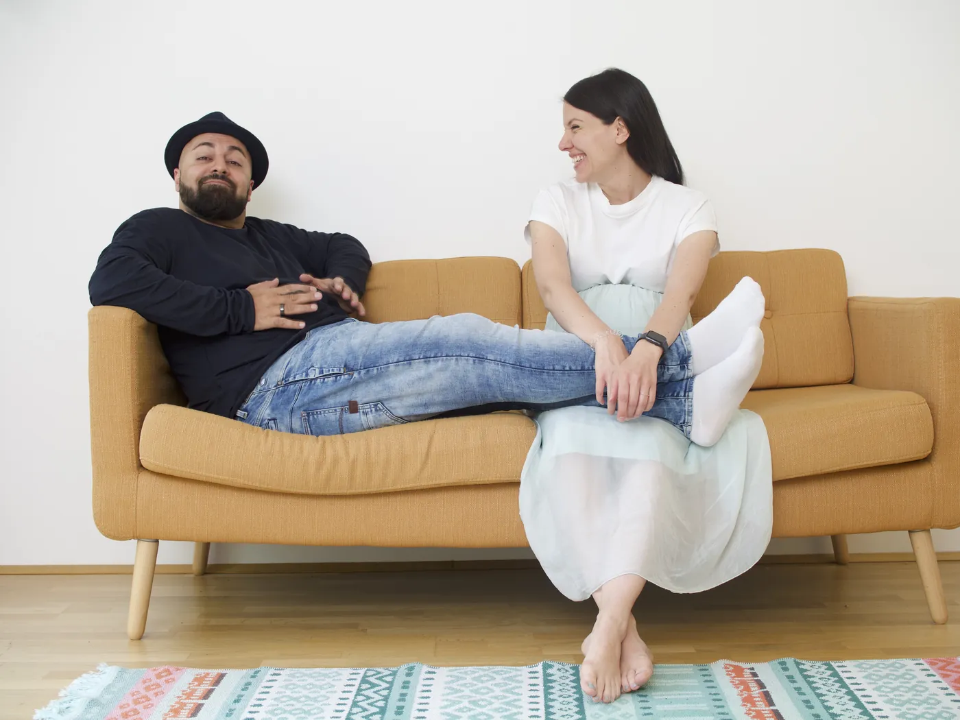 Ali mit Hut lehnt auf einem gelben Sofa, die Beine auf Annas Schoß, beide lachen