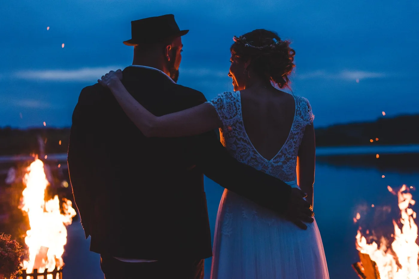 Anna und Ali Mahlodji am Hochzeitstag, von hinten, Arm in Arm am See in der blauen Stunde neben einem Feuer