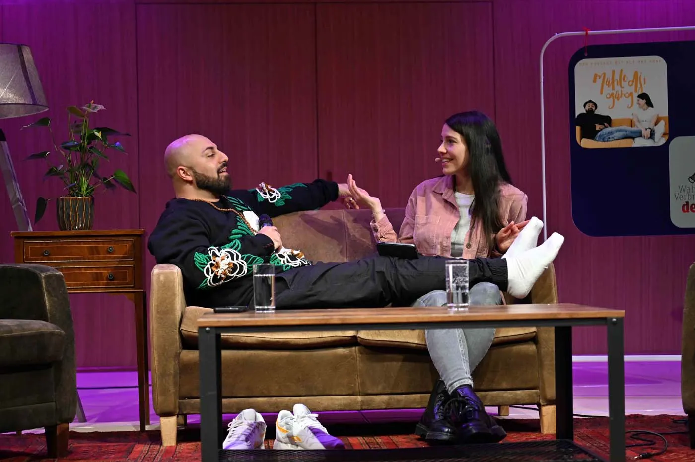 Anna und Ali auf einer Bühnen-Couch, Ali hat die Beine hochgelegt, lila Bühnenlicht