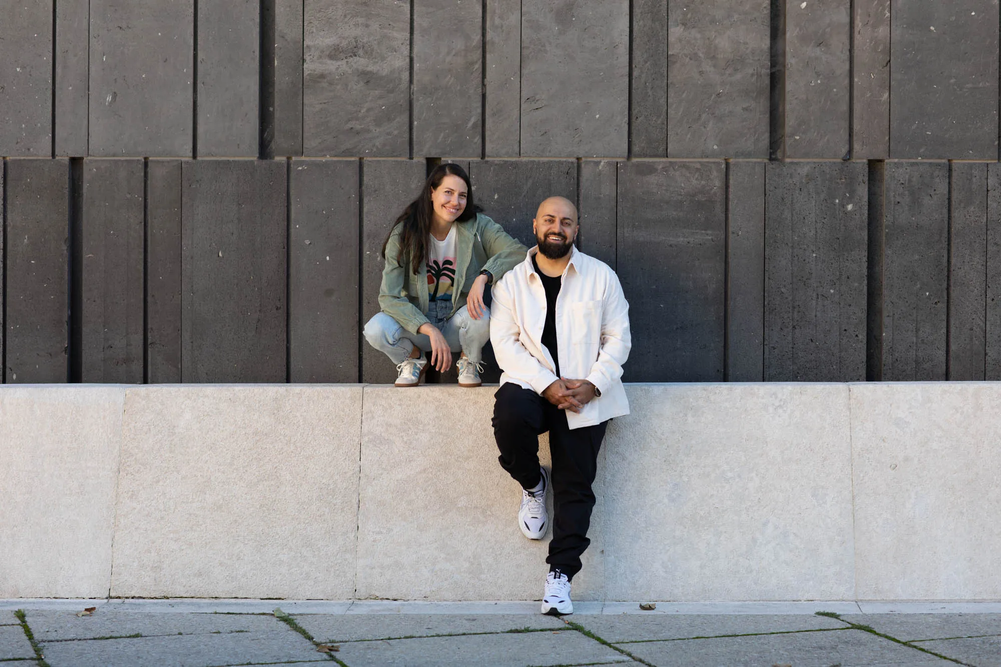 Anna and Ali Mahlodji on a stone wall in front of a dark facade: she crouches on the wall laughing, he sits in front of it