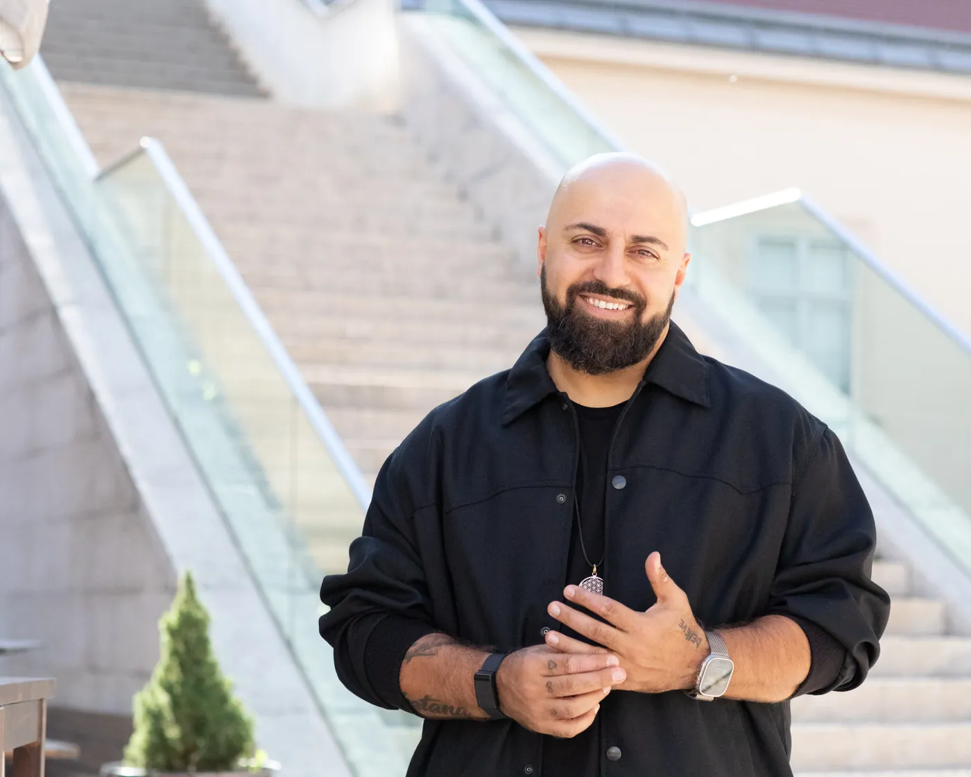 Ali Mahlodji lächelnd vor einer Treppe, die Hände vor der Brust