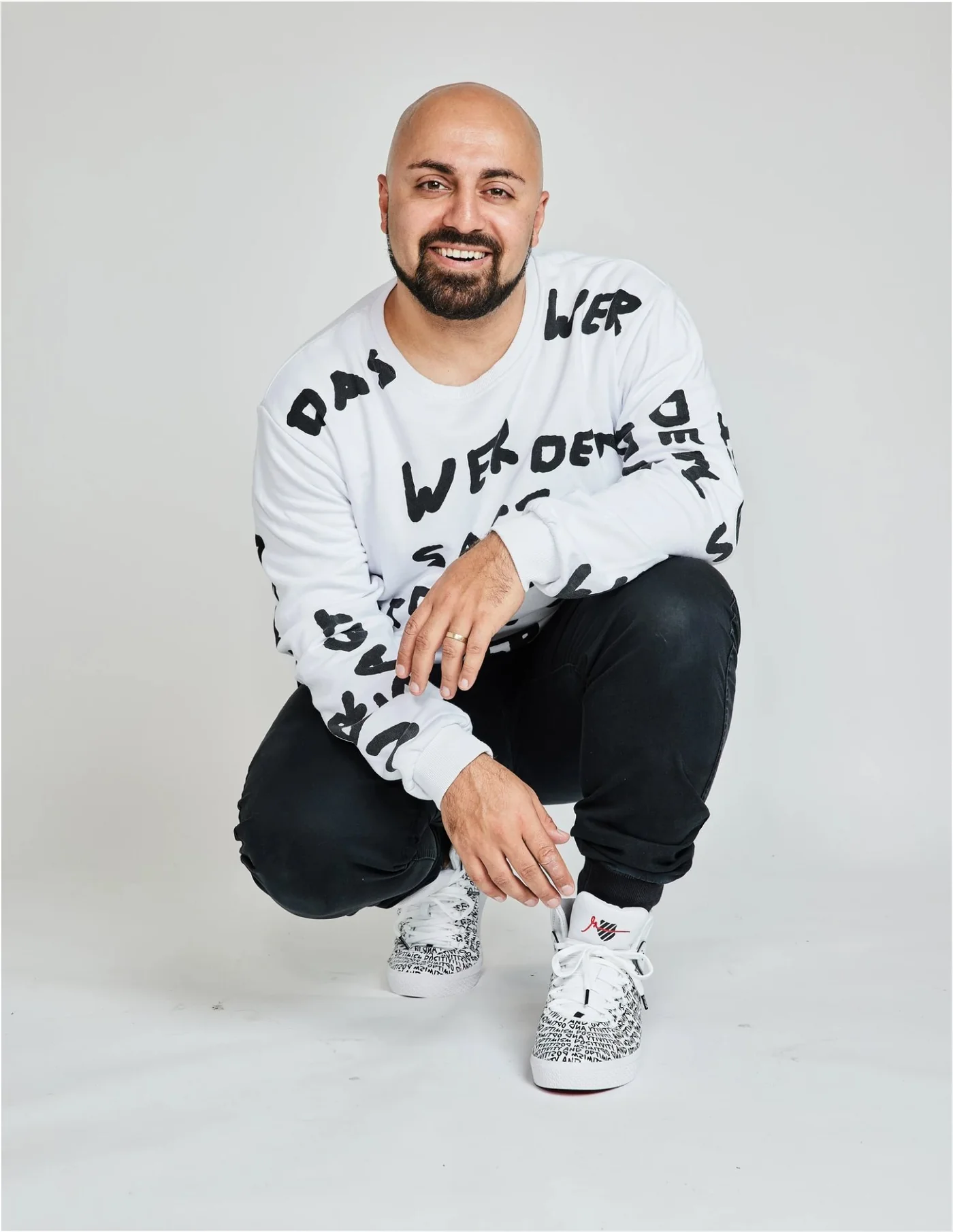 Ali Mahlodji in der Hocke, Sweater mit Schriftzug „Das, wer, dem, sag, bist, wie …“